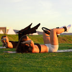 Two women in sportswear exercise on mats at sunset, using SMAI Weighted Ankle/Wrist Bracelets 1KG (Pair) - Black and Pilates rings for resistance training outdoors on a grassy field.