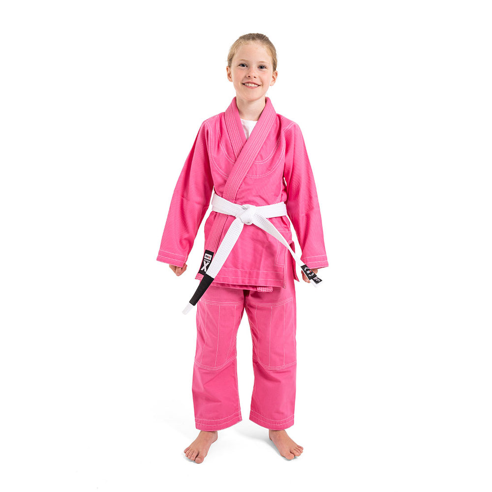 A smiling child in a SMAI Supreme X Brazilian Jiu Jitsu Uniform - Pink and a white belt stands barefoot against a plain white background.