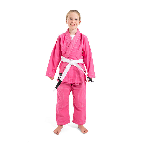 A smiling child in a SMAI Supreme X Brazilian Jiu Jitsu Uniform - Pink and a white belt stands barefoot against a plain white background.