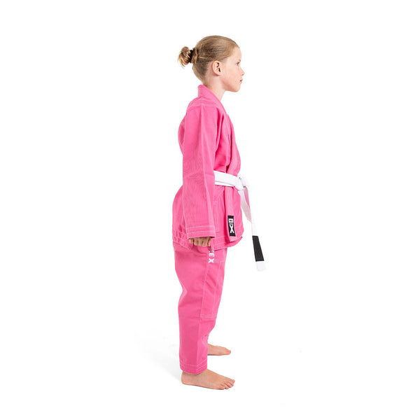 A young child stands in profile against a white background, wearing the SMAI Supreme X Brazilian Jiu Jitsu Uniform in pink, crafted from durable Rip Stop fabric and finished with a white belt tied at the waist.