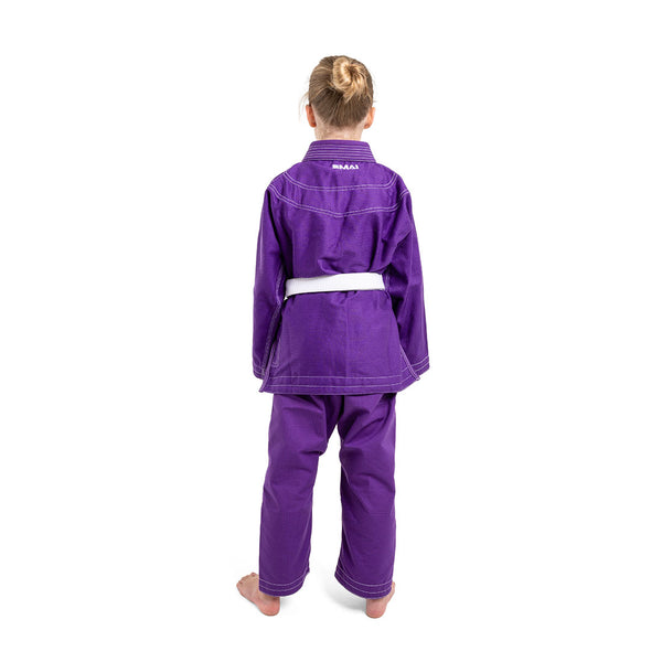 A young child with blonde hair in a bun wears the SMAI Supreme X Brazilian Jiu Jitsu Uniform in purple, featuring pearl weave fabric and Rip Stop pants, standing barefoot facing away against a white background.