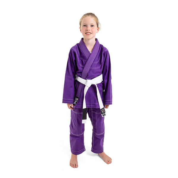 A young child stands barefoot and smiling in the SMAI Supreme X Brazilian Jiu Jitsu Uniform – Purple, featuring pearl weave fabric and a white belt, against a plain white background.