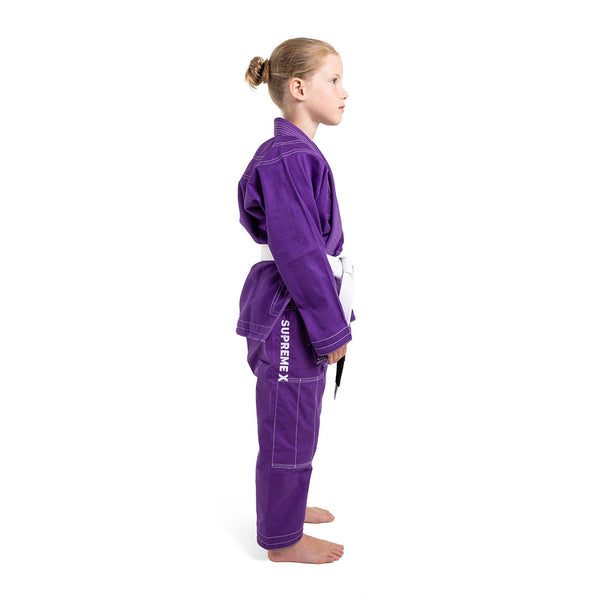A young child stands in profile, barefoot against a white background, wearing the SMAI Supreme X Brazilian Jiu Jitsu Uniform in purple with pearl weave fabric, Rip Stop pants labeled "SUPREME X," and a white belt.