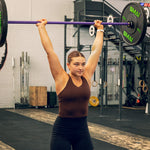 A focused woman in athletic wear lifts the SMAI Barbell - Purple - 20kg overhead at the gym, with fitness equipment and weightlifting racks around her. Her hair is tied back.