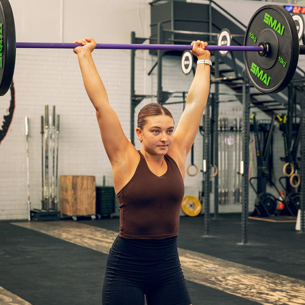 A focused woman in athletic wear lifts the SMAI Barbell - Purple - 20kg overhead at the gym, with fitness equipment and weightlifting racks around her. Her hair is tied back.
