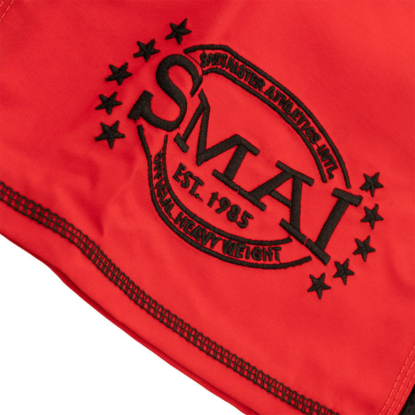 Close-up of red heavy-duty MMA Shorts by SMAI, featuring black embroidery with "SMAI EST. 1985," "Universal Heavy Weight," and "Sports Master Athletics Intl" in an oval design, highlighting durability for intense training.