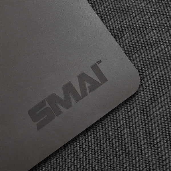 A close-up of the SMAI Pilates Reformer Carriage Topper / Protector, a gray rectangular mat with “SMAI” printed in dark letters, shown on a textured dark gray fabric surface.