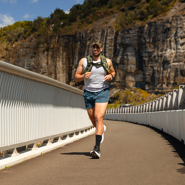 A man wearing the SMAI Weighted Ruck Vest - Pink runs across a curved bridge in a cap, tank top, and shorts, with rocky cliffs and greenery under a blue sky.