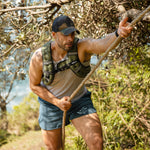 A man wearing a cap, tank top, shorts, and the SMAI Weighted Ruck Vest - Pink climbs a rope outdoors among trees and greenery.