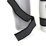 A Taekwondo Shin Guard by SMAI is showcased against a white background, featuring a black and white striped elastic closure. It is crafted with lightweight carbon fibre to ensure optimal protection. Part of another object can be seen partially on the right side.