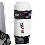 The Taekwondo Shin Guard by SMAI is designed in white with black and white adjustable straps, featuring EVA foam padding. The brand name "SMAI" is printed in red and black, and its elastic closure ensures a secure fit, making it perfect for martial arts or sports use.