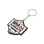 The SMAI Karate Keyring - Rubber features the rectangular "SMAI KARATE" logo in black and white with a red "A," a stylized fist outline in the background, and comes attached to a chain and keyring.