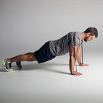 A man is in a plank position, using the SMAI Rubber Resistance Band - 100lb looped around his shoulders with his hands on the floor. He sports a gray shirt, black shorts, and lime green sneakers against a plain white background.
