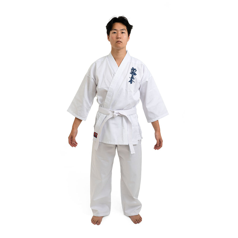 On a white background, a person stands barefoot, wearing the SMAI Shinkyokushinkai Uniform - 8oz Student Gi with a belt. Dark lettering adorns the left side of the top. With arms relaxed at their sides and facing forward, they encapsulate Shinkyokushinkai discipline.