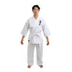 On a white background, a person stands barefoot, wearing the SMAI Shinkyokushinkai Uniform - 8oz Student Gi with a belt. Dark lettering adorns the left side of the top. With arms relaxed at their sides and facing forward, they encapsulate Shinkyokushinkai discipline.