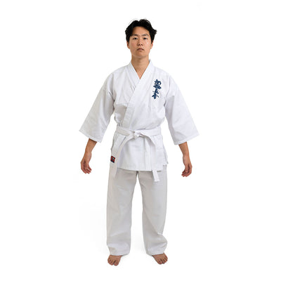On a white background, a person stands barefoot, wearing the SMAI Shinkyokushinkai Uniform - 8oz Student Gi with a belt. Dark lettering adorns the left side of the top. With arms relaxed at their sides and facing forward, they encapsulate Shinkyokushinkai discipline.