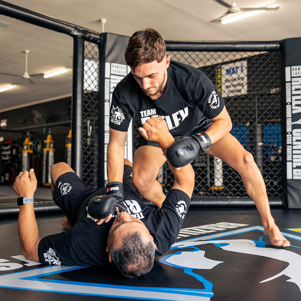 Inside an octagon training cage, a mixed martial arts fighter straddles and pins down an opponent during a sparring session. Both wear black shirts and SMAI Elite85 MMA Hybrid Sparring Gloves.