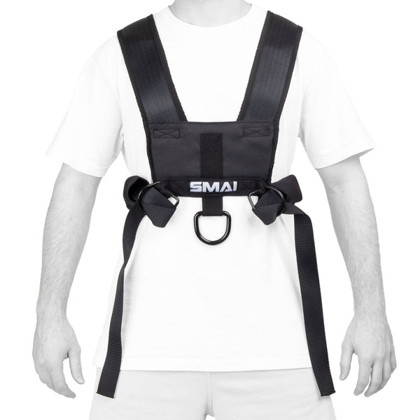 A person wears the SMAI Tower Gym Sled 3.0 with sled harness—featuring wide black straps and a front metal ring—over a white t-shirt and light shorts, preparing for sprint training.