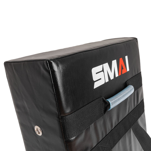 The SMAI Kick Shield - Shoc Tec Straight is a black rectangular pad with a handle and straps, made from lightweight materials and impact-absorbing foam for safe martial arts training. "SMAI" is printed on the surface.
