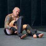 A tattooed man in MMA gloves and shorts sits on a mat in a dark-walled gym, putting on SMAI Elite85 Muay Thai Shin Guards - Black.