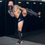 A muscular, tattooed man practices a high kick in a gym, wearing SMAI Elite85 Muay Thai Shin Guards in black along with MMA gear, focused and poised in a fighting stance against padded black walls and mats.