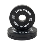 SMAI's The Yard - 7.5kg Fractional Plate Set features black Olympic barbell plates with white “THE YARD EST 2020 1.25 KG 2.5 LB” text—perfect for micro-loading, with one plate upright and others stacked flat for organized workouts.