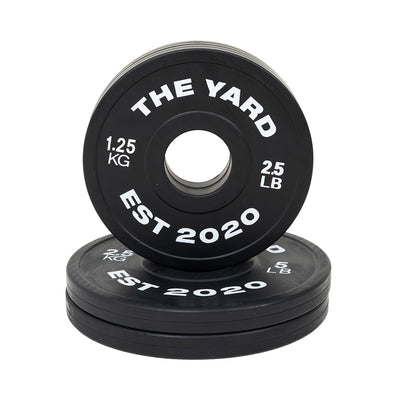 SMAI's The Yard - 7.5kg Fractional Plate Set features black Olympic barbell plates with white “THE YARD EST 2020 1.25 KG 2.5 LB” text—perfect for micro-loading, with one plate upright and others stacked flat for organized workouts.