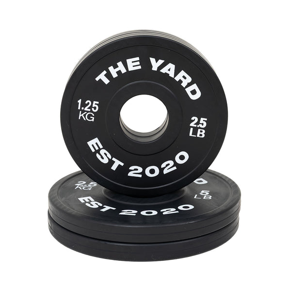 SMAI's The Yard - 7.5kg Fractional Plate Set features black Olympic barbell plates with white “THE YARD EST 2020 1.25 KG 2.5 LB” text—perfect for micro-loading, with one plate upright and others stacked flat for organized workouts.