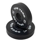 Two black SMAI Olympic plates from The Yard - 7.5kg Fractional Plate Set show "EST 2020" in white. One upright plate shows "1.25 KG" and "2.5 LB," others display "5 LB." Perfect for micro-loading and incremental progress.