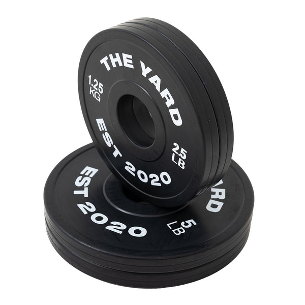 Two black SMAI Olympic plates from The Yard - 7.5kg Fractional Plate Set show "EST 2020" in white. One upright plate shows "1.25 KG" and "2.5 LB," others display "5 LB." Perfect for micro-loading and incremental progress.