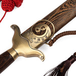 Close-up of the SMAI Tai Chi Sword Brass Fitting Ying Yang, featuring a brass guard engraved with a yin-yang symbol and Chinese characters, attached to a wooden sheath with brown tassel and red braided cord.