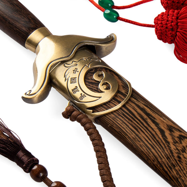 Close-up of the SMAI Tai Chi Sword Brass Fitting Ying Yang, featuring an ornate brass guard with engraved Yin-Yang symbol, spring steel blade, dark wooden scabbard, and decorative red and brown tassels.