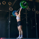 Dressed in athletic wear, a person lifts a barbell overhead, demonstrating power and focus amidst gymnastic rings and SMAI's Competition Bumper Plate 20kg (PAIR) IWF Colour plates at the gym.
