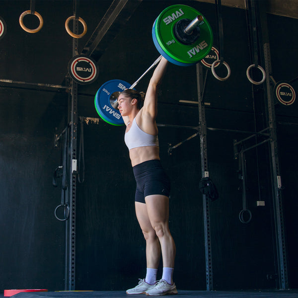 In a lively gym, an individual in a white sports bra and black shorts expertly hoists a barbell with SMAI Competition Bumper Plates (10kg, IWF Colour) overhead amid hanging gymnastic rings, showcasing the essence of weightlifting contests.