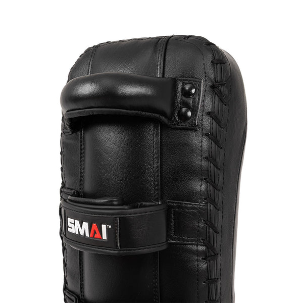 A close-up of the SMAI Muay Thai Pad - Syntec Black (Pair) shows black synthetic leather, visible stitching, a top handle, and a lower strap with the SMAI logo.