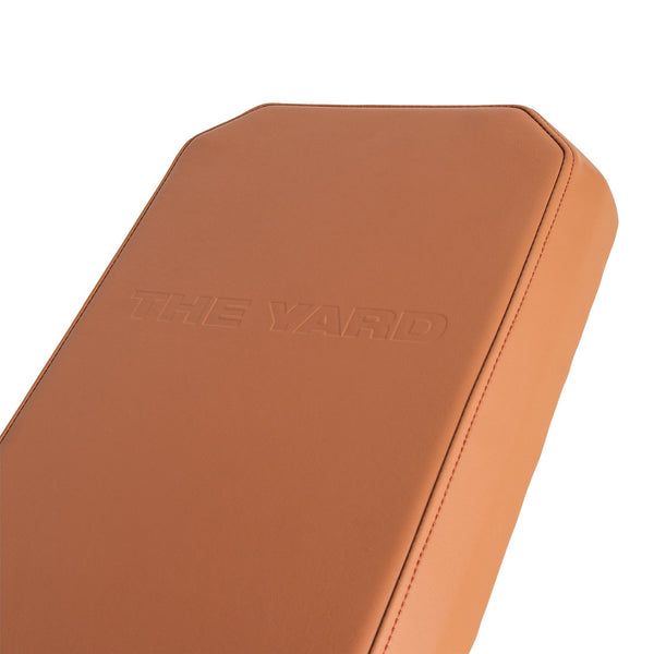 A close-up of the tan leather cushion on the SMAI Yard Adjustable Weight Bench - Super Bench shows "THE YARD" embossed on its surface with neat stitching along the edges.