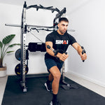 A man in a black SMAI t-shirt and shorts performs a standing cable chest fly using the SMAI All in 1 Functional Trainer in his home gym, with a plant and weights visible in the background.