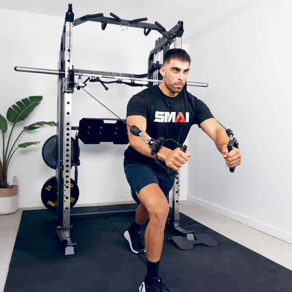 A man in a black SMAI t-shirt and shorts performs a standing cable chest fly using the SMAI All in 1 Functional Trainer in his home gym, with a plant and weights visible in the background.