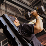 A person in a black shirt with a tattoo lifts SMAI Rubber Hex Dumbbells Classic (Pair) with an ergonomic grip while seated on an inclined bench in a commercial gym with industrial-style ceilings.