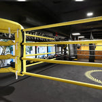 The 5m Boxing Ring - Competition by SMAI features bright yellow ropes, a high gauge steel frame, a black mat with a laurel design at the center, and is encircled by punching bags and windows in the gym's free-standing, well-lit space.