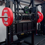 A Vanta Series Leg Press Attachment by SMAI is set up in a home gym, with red 25kg SMAI plates on the barbell, extra weights stored on the rack, and multiple dumbbells visible in the background.