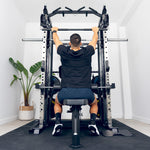 A person sits on a bench in a home gym, using the SMAI All in 1 Functional Trainer for a lat pulldown. The gym has white walls and a potted plant in the corner.