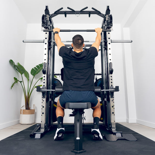 A person sits on a bench in a home gym, using the SMAI All in 1 Functional Trainer for a lat pulldown. The gym has white walls and a potted plant in the corner.