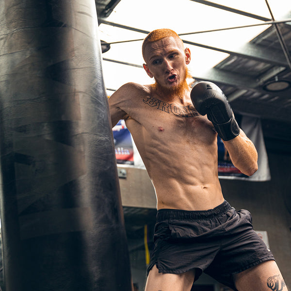 A shirtless man with red hair and a beard, wearing black boxing gloves and shorts, punches the SMAI Punching Bag - 5ft Triple Black in a gym with a glass ceiling.