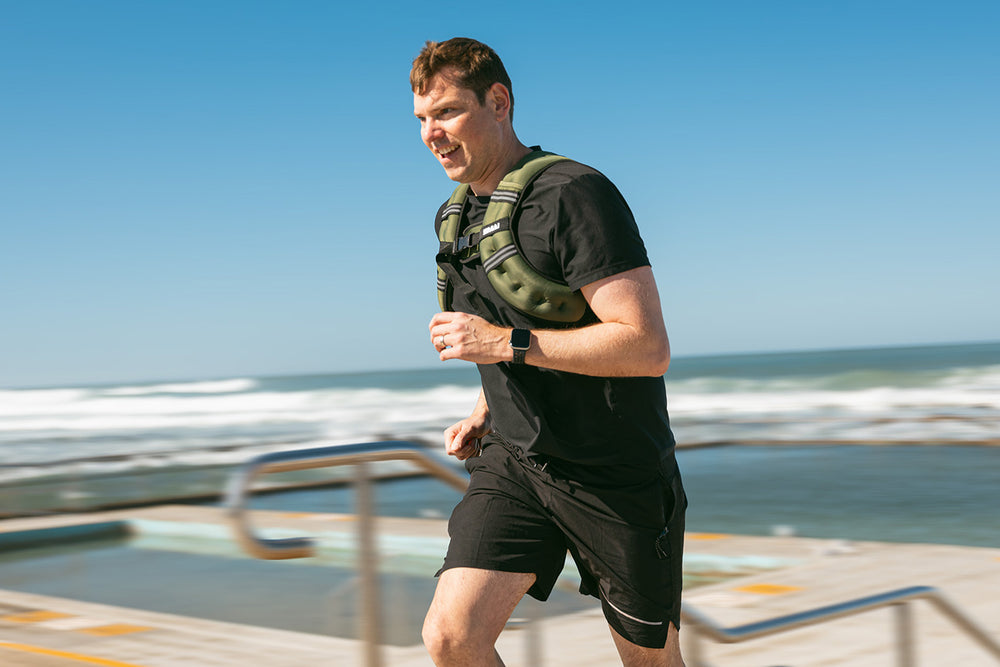 A man in black athletic wear runs outdoors near the ocean, wearing a green weighted vest. The sky is clear and the waves can be seen in the background.