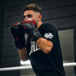 A young man in a black T-shirt stands in a boxing stance at the gym, holding up SMAI's Black Dragon Limited Edition Boxing Gloves.