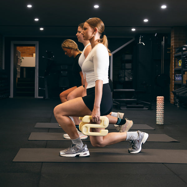 Three women exercise indoors at a gym, performing lunges on mats. The woman in front uses the SMAI SBELL Starter pack, holding weights in each hand. All wear athletic gear and sneakers, with gym equipment and dim lighting in the background.