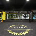 A 6m SMAI Boxing Ring with black and yellow ropes is pictured. The floor is black with a laurel wreath design and "Olympia" text. Boxing gloves adorn the ropes, and a digital clock shows 2:36. Large windows reveal trees outside.