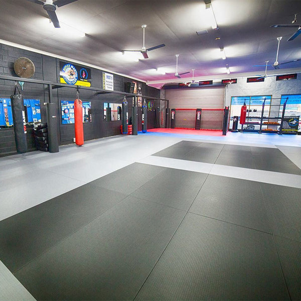 A spacious martial arts gym features SMAI Judo/BJJ Mats for flooring, padded walls, punching bags, wall mirrors, and large windows for natural light. Signs and organized equipment line the clean black walls.