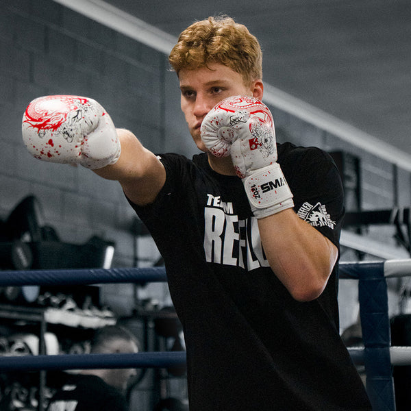 A young man with curly hair, wearing SMAI Boxing Glove - White Dragon Limited Edition and a black T-shirt, throws a punch in a gym boxing ring. Focused, he trains hard.